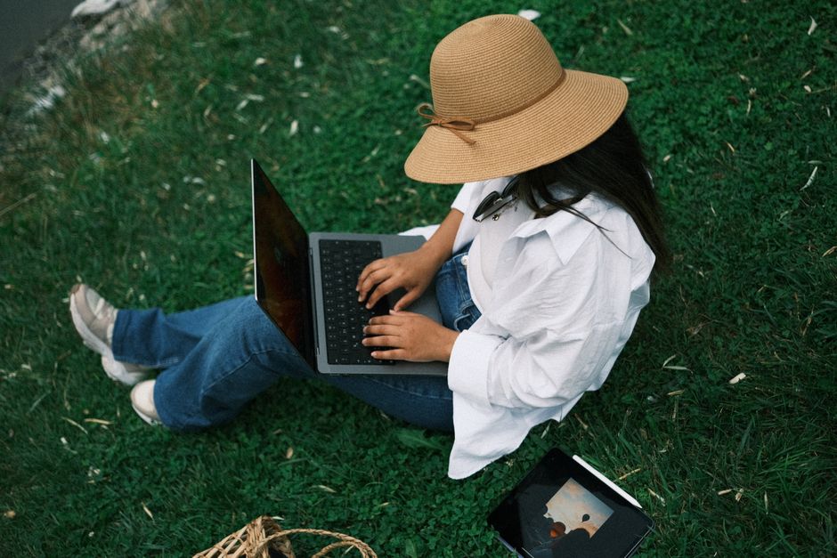 A woman in a hat works on her laptop outdoors, sitting on grass in a park setting.