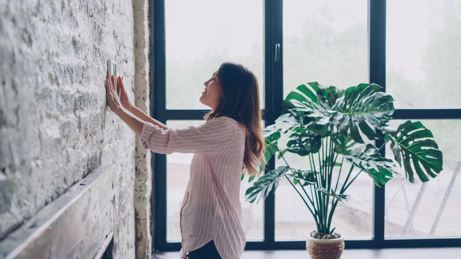 Woman adjusting the thermostat in a bright, contemporary room with large windows and a potted plant.